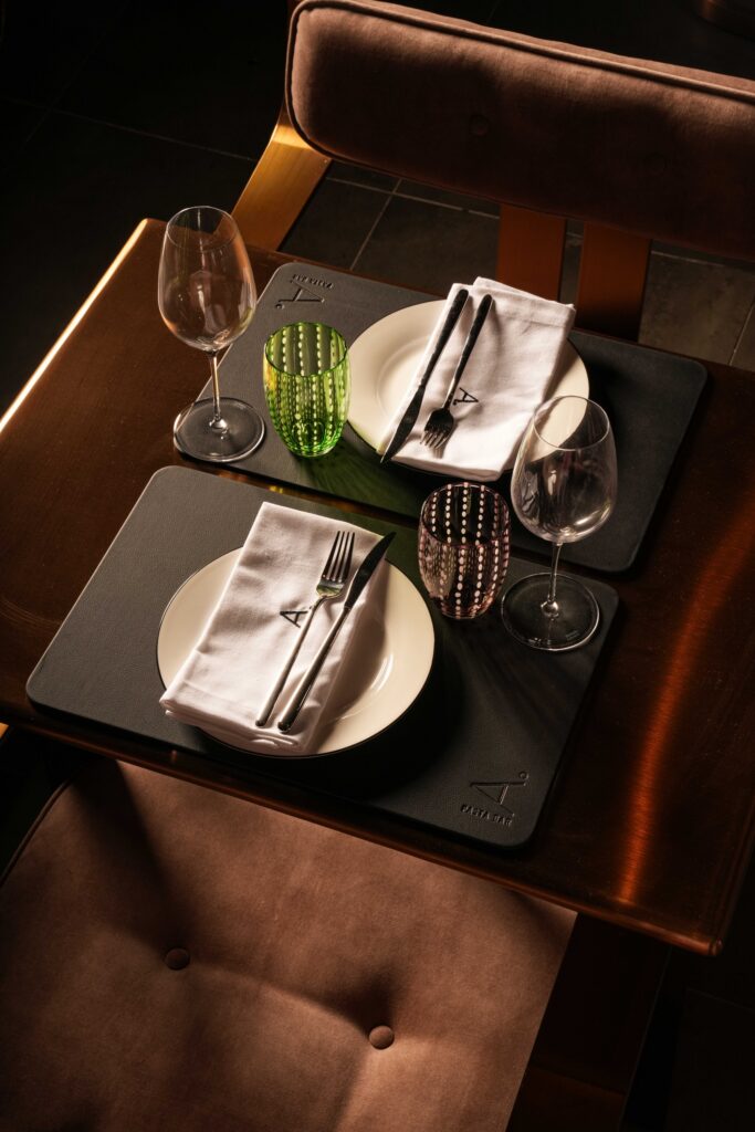 Restaurant table for two with white plates, colourful glasses and brown upholstered chairs.