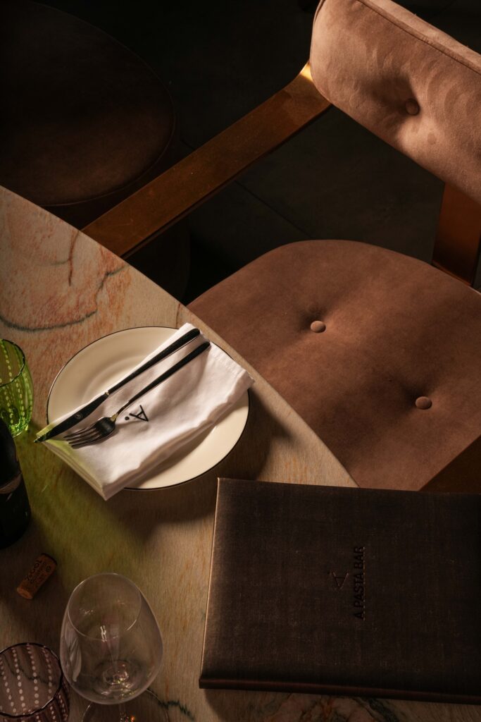Sunlit place setting and brown menu beside an upholstered restaurant chair.