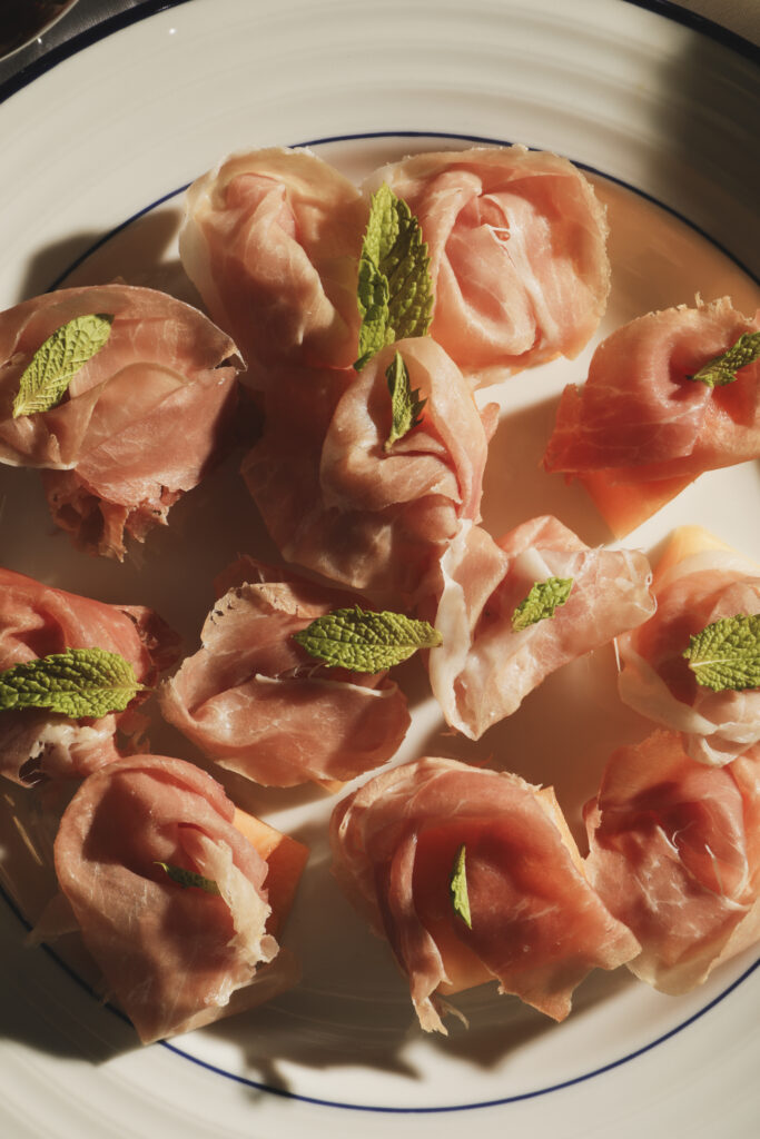 Prosciutto and melon topped with mint leaves on a white plate.