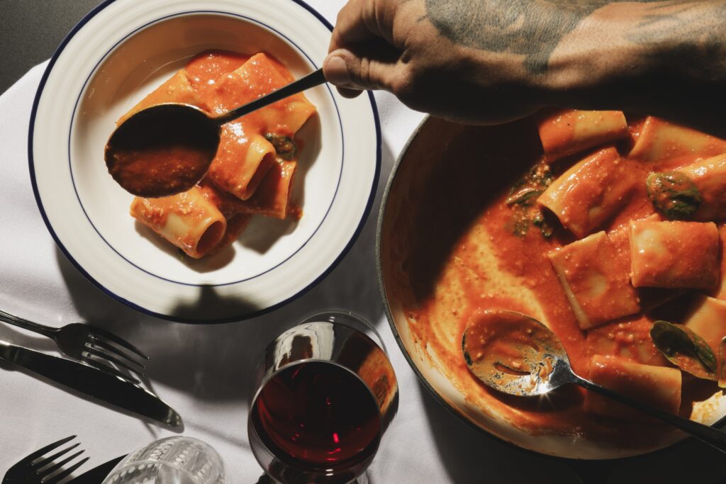 A diner serving pasta tubes in tomato sauce from a large bowl onto a plate.