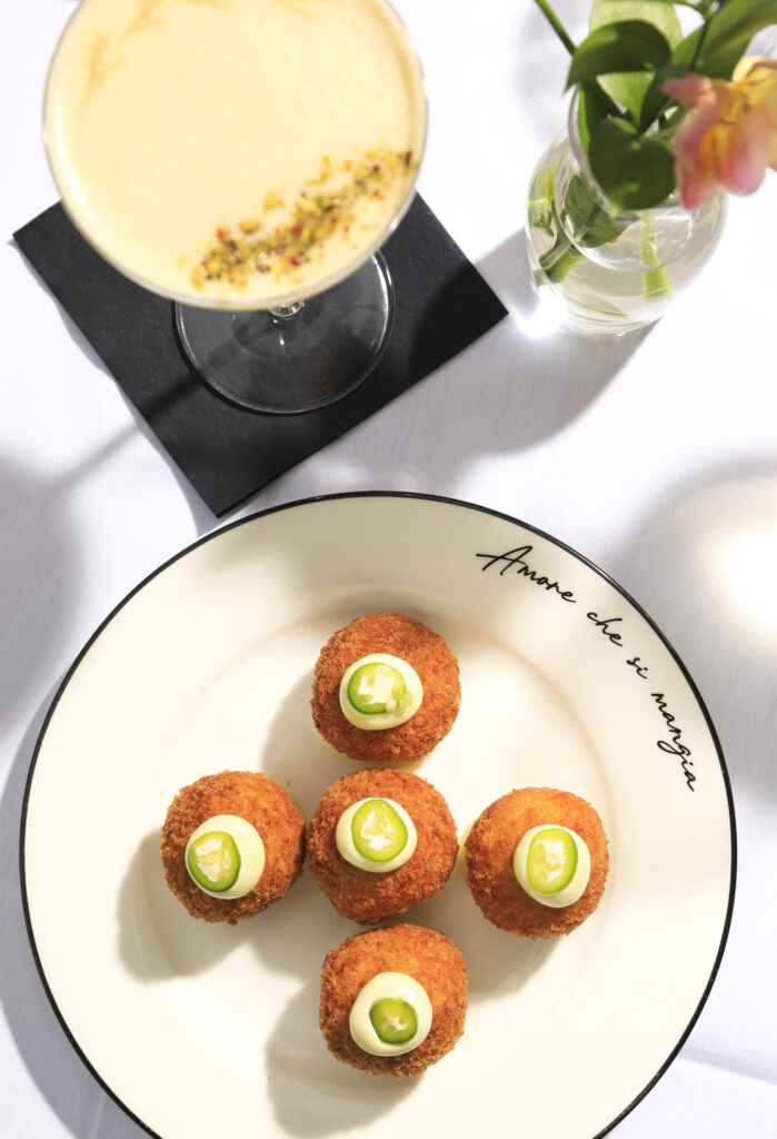 Five golden croquettes with sauce and green chilli slices, served beside a yellow cocktail.