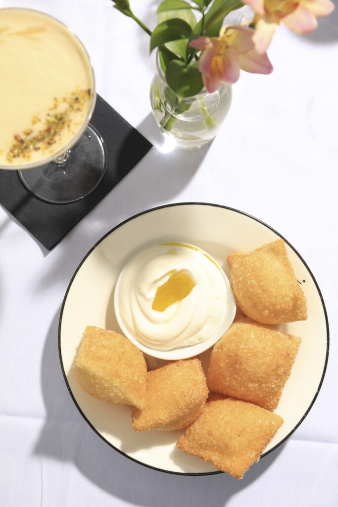 Golden fried dough squares served with a bowl of cream beside a yellow cocktail.