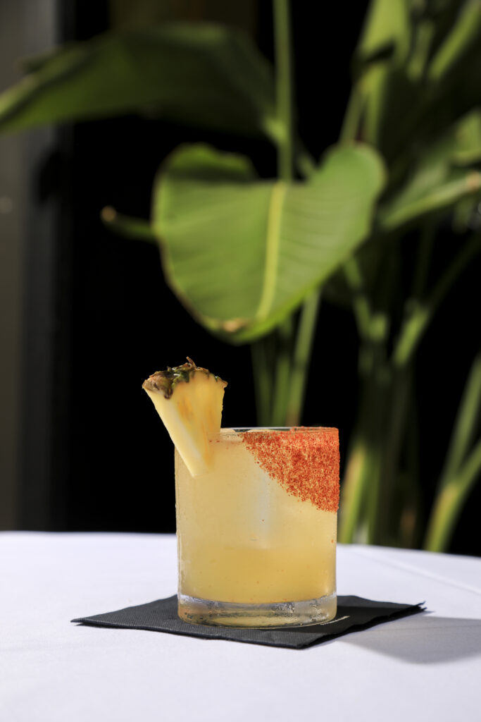 Yellow cocktail with a pineapple wedge and a red seasoned rim.