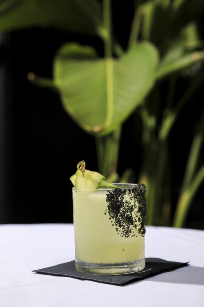 Pale green cocktail with cucumber ribbons and a dark seasoned rim.