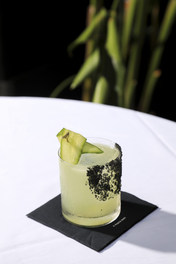 Cucumber-garnished green cocktail on a black napkin in the sunshine.