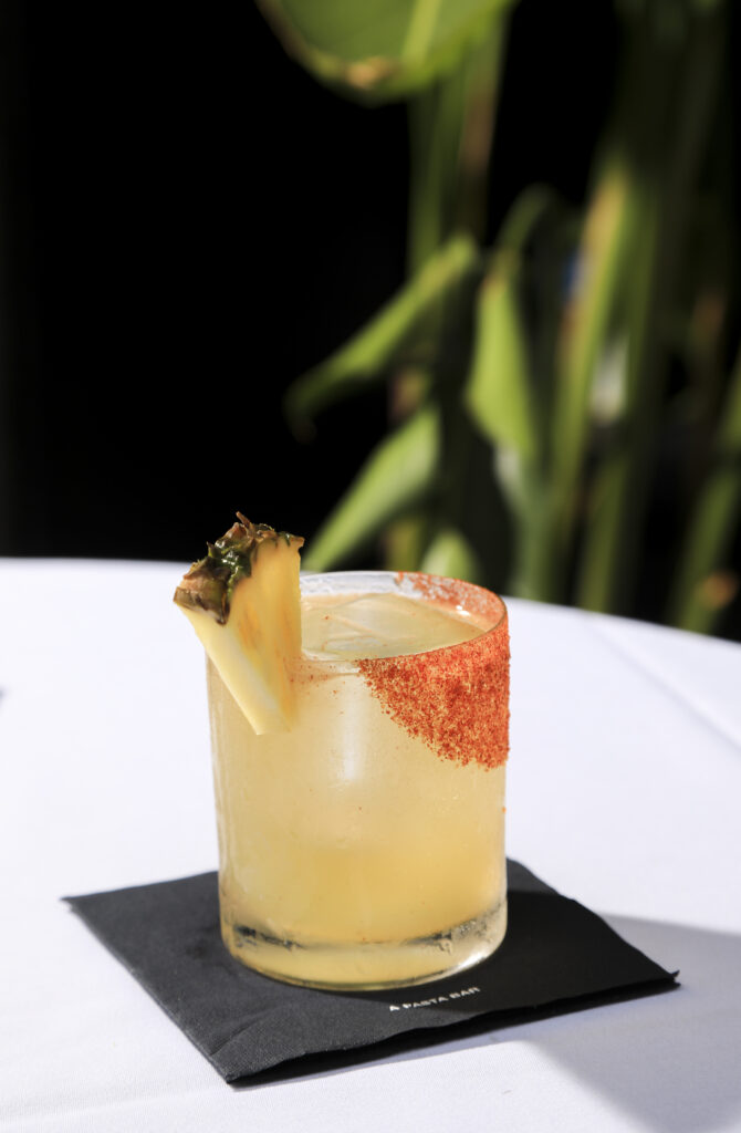 Pineapple-garnished yellow cocktail with a red seasoned rim on a white table.
