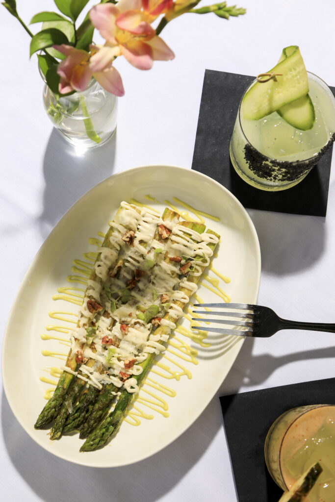 Grilled asparagus drizzled with creamy dressing beside a cucumber cocktail.