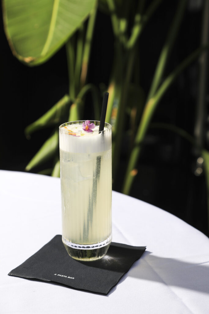 Tall pale green cocktail with a foamy top, a straw and a small flower garnish.