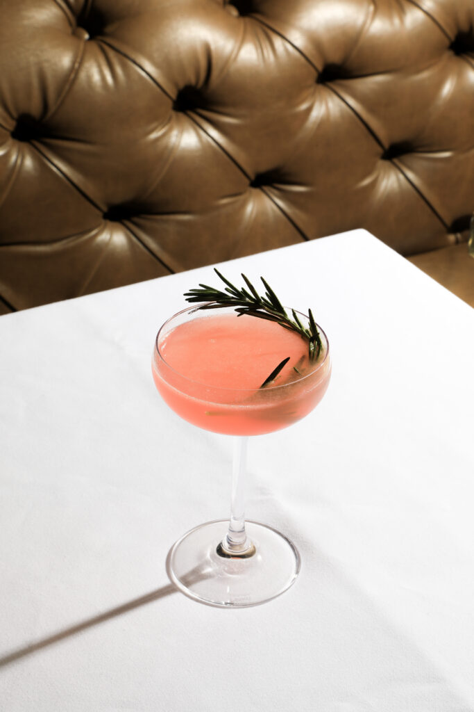 A pink cocktail garnished with a sprig of rosemary on a white tablecloth.