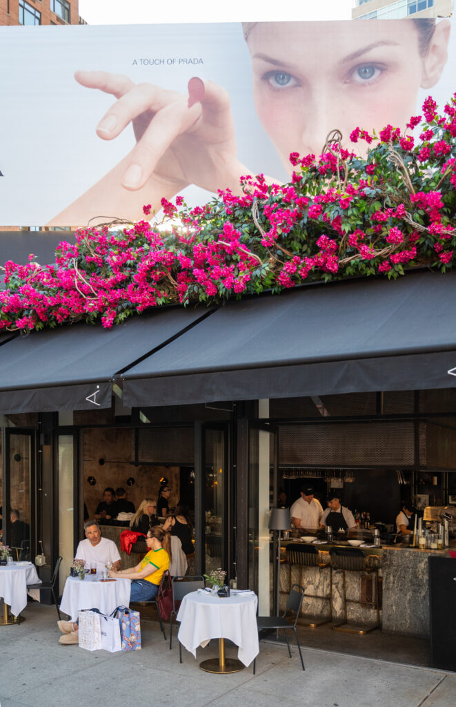 Outdoor diners beneath a black awning and bright pink flowers at the restaurant entrance.