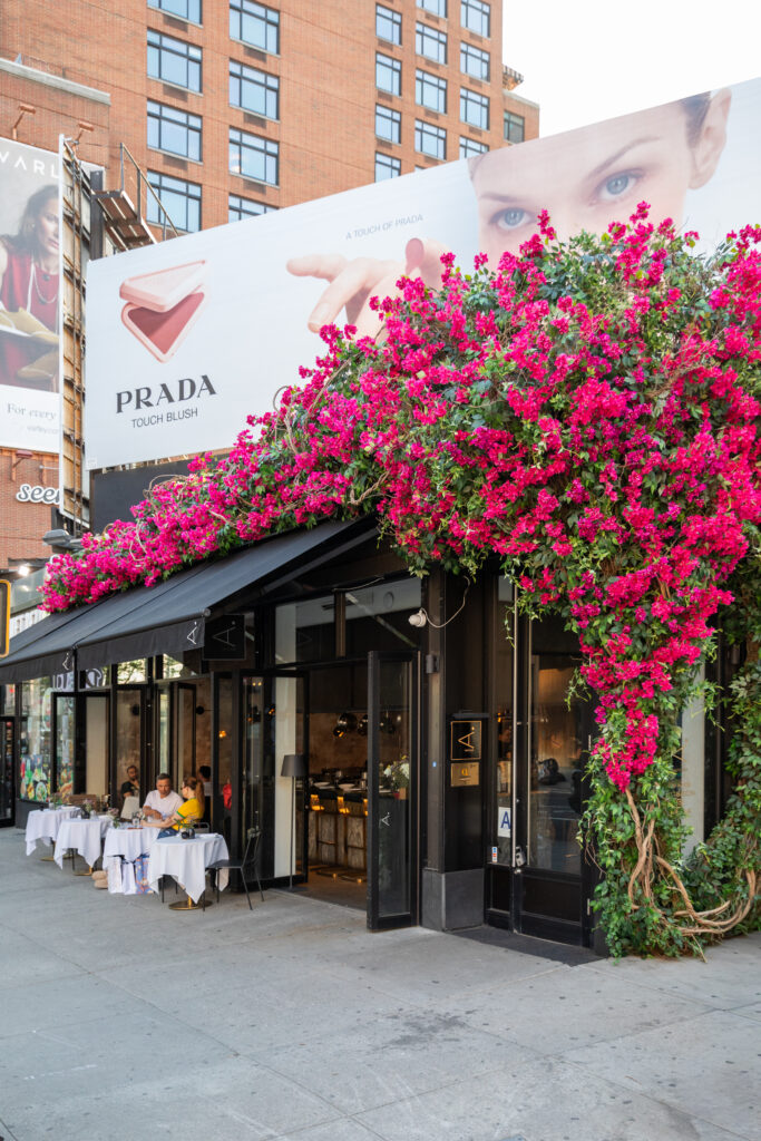 The restaurant's corner entrance framed by pink flowers, with pavement dining beneath a black awning.