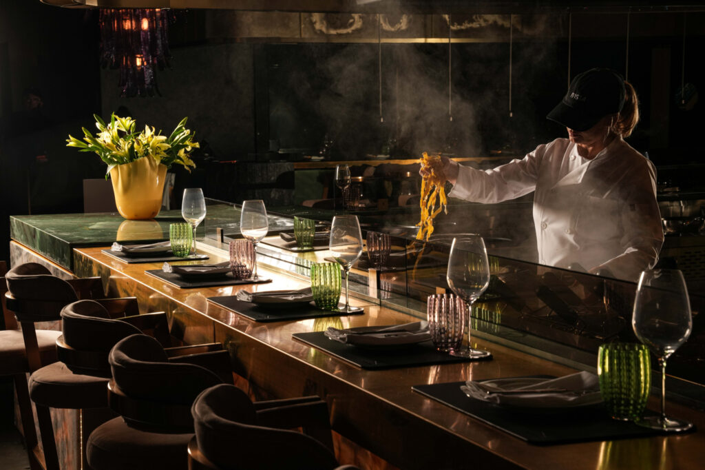 Chef lifting long strands of pasta above a steaming pan in the open kitchen.