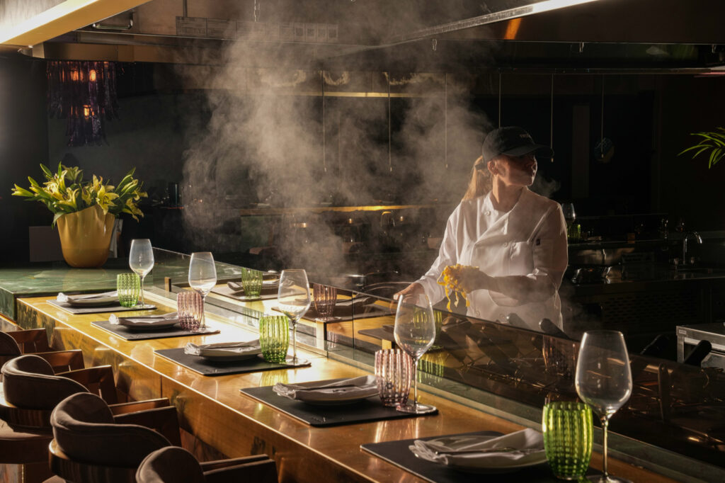 Chef lifting pasta above a steaming cooking station behind the dining counter.