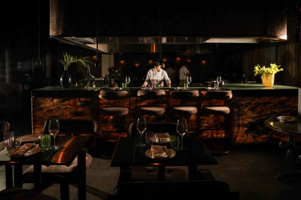 Chefs working behind an illuminated kitchen counter, with set dining tables in the foreground.