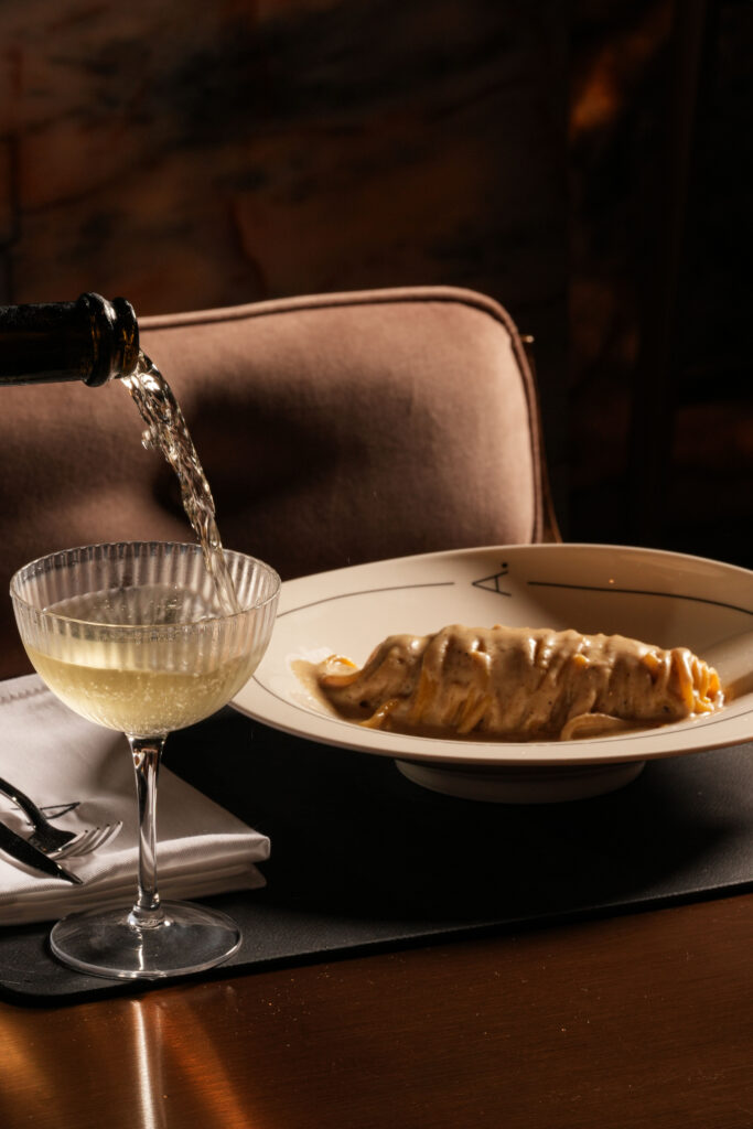 White wine being poured into a glass beside a plate of pasta.