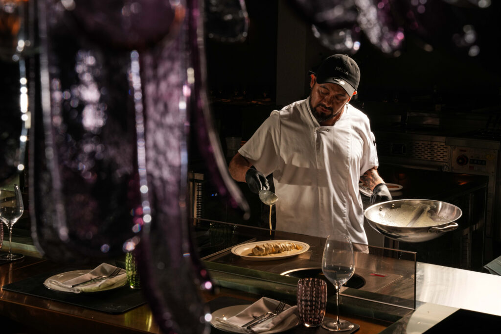 Chef spooning sauce over plated pasta while holding a pan in the open kitchen.