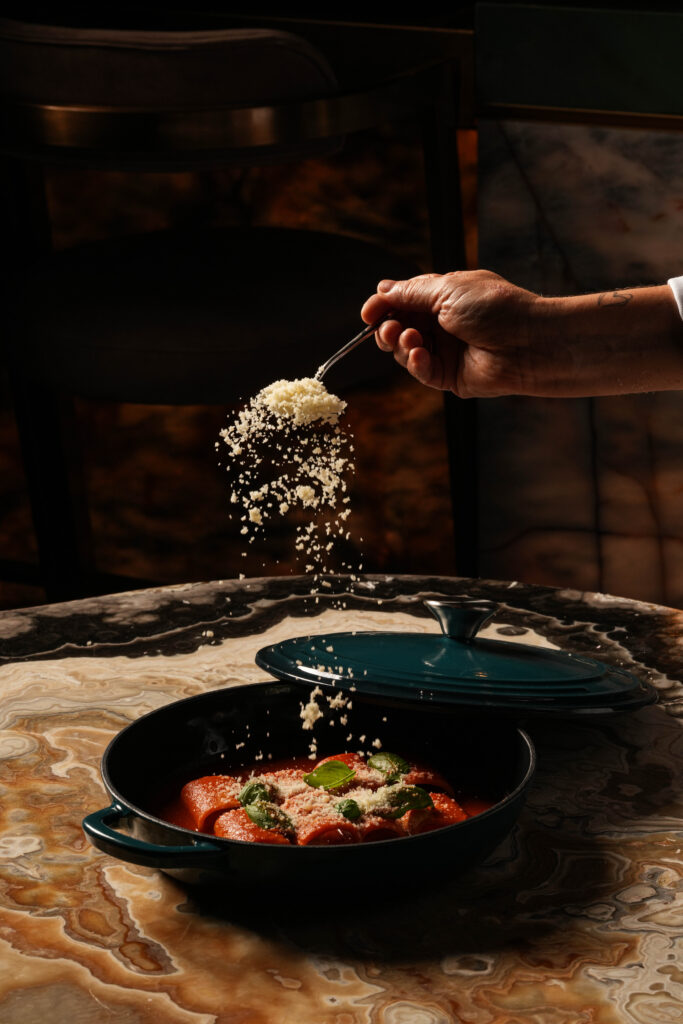 Grated cheese being sprinkled over pasta in tomato sauce with basil in a black pan.