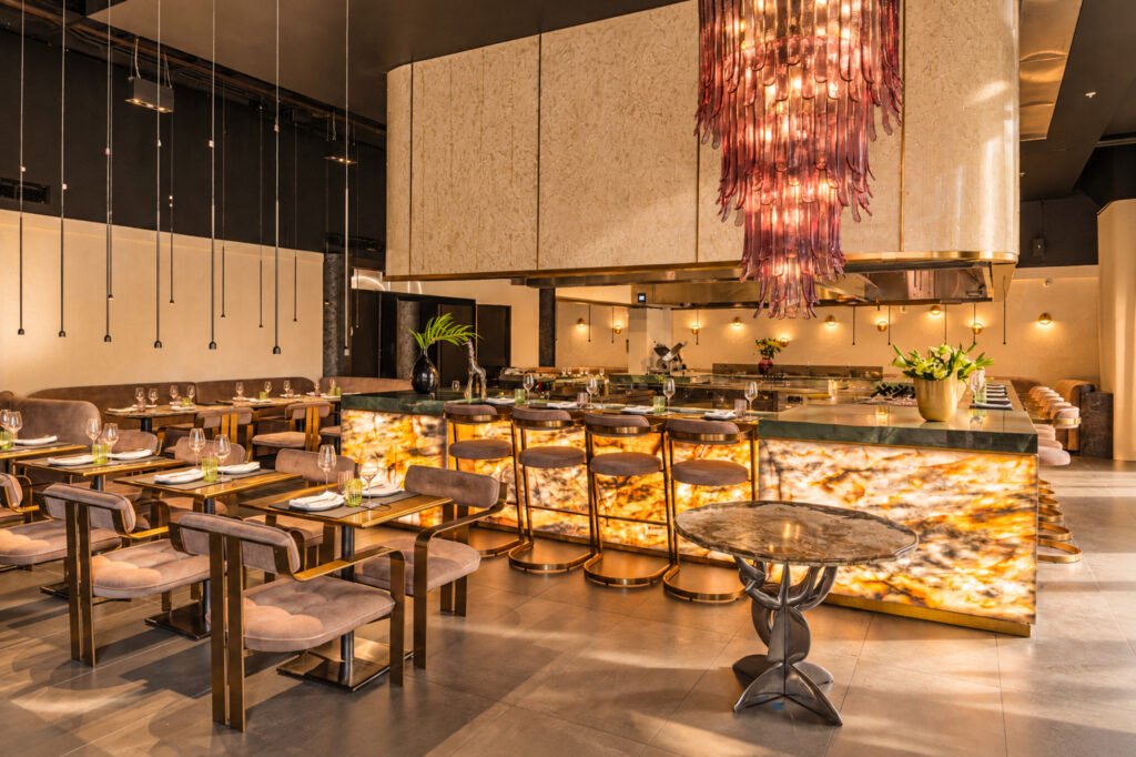 Dining tables and counter seating around an open kitchen beneath a pink glass chandelier.