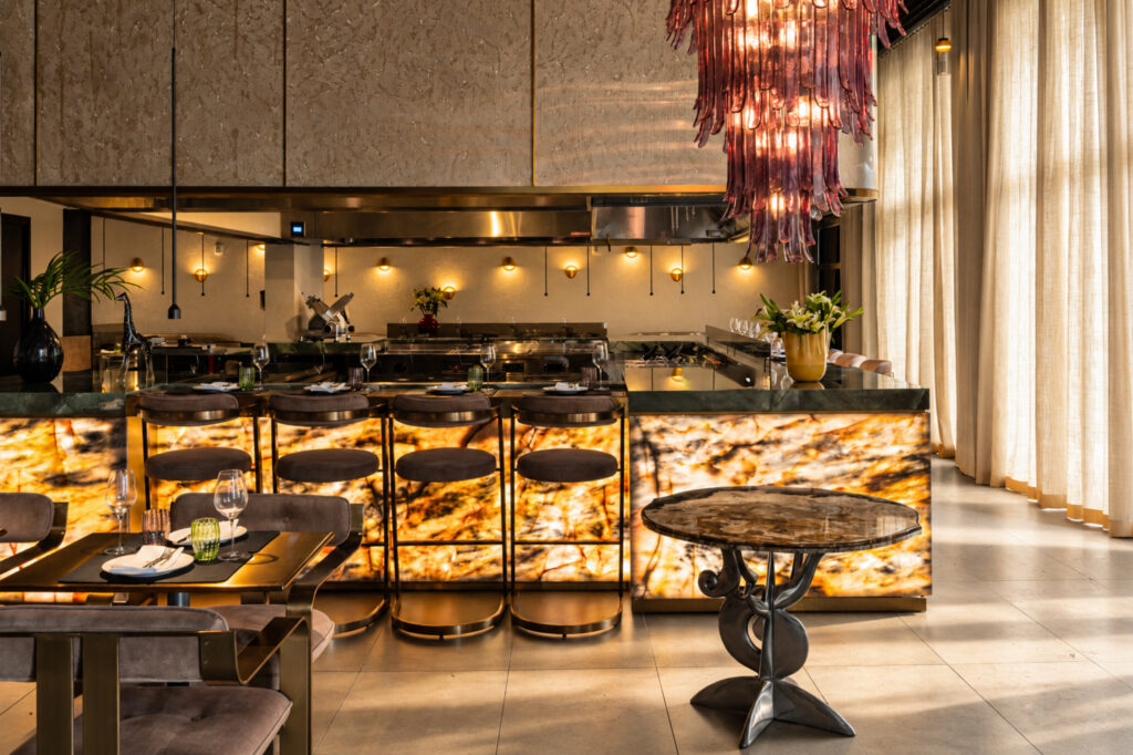 Illuminated stone kitchen counter with stools beneath a pink glass chandelier.